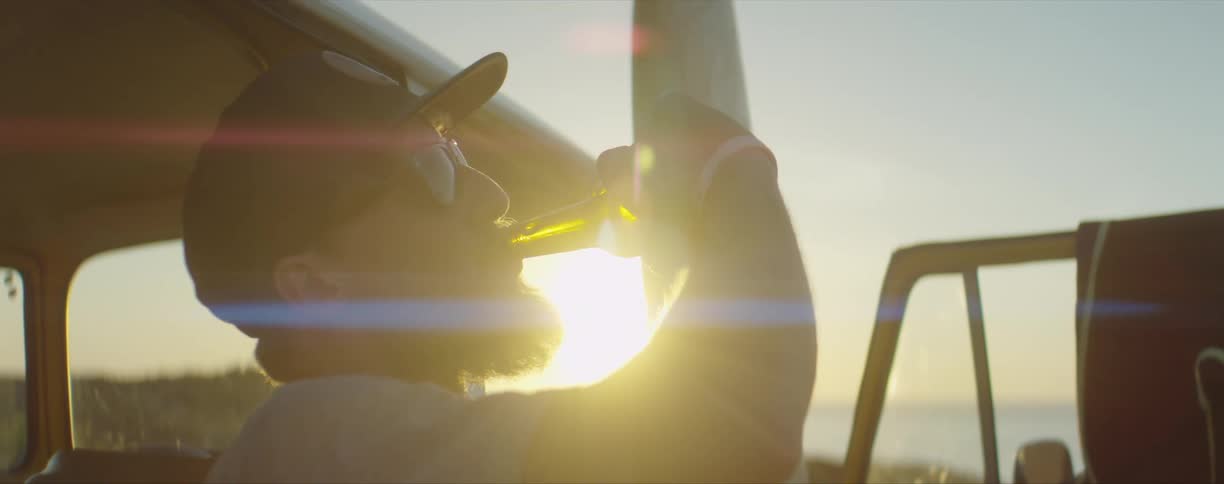 A man with a hat and sunglasses drinking a beer from the bottle with the sunset the sky and clouds in the background