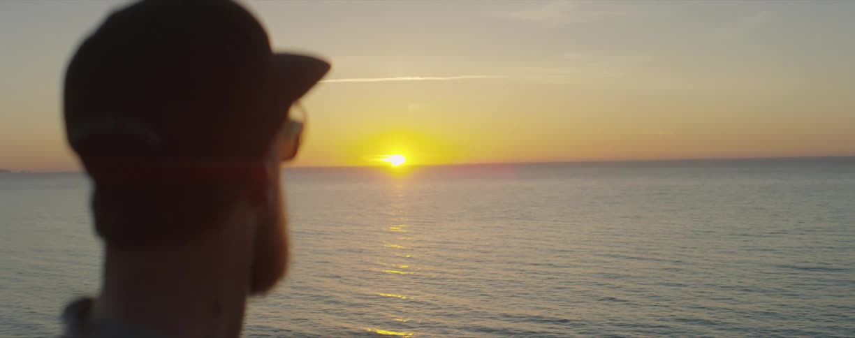 Man wearing sunglasses looking at the sun setting on the horizon over the ocean