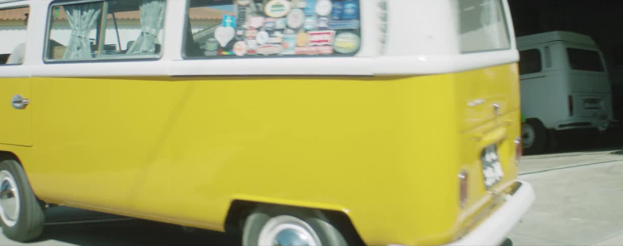 A caucasian man driving a volkswagen van up an alley around a building to other parked vans