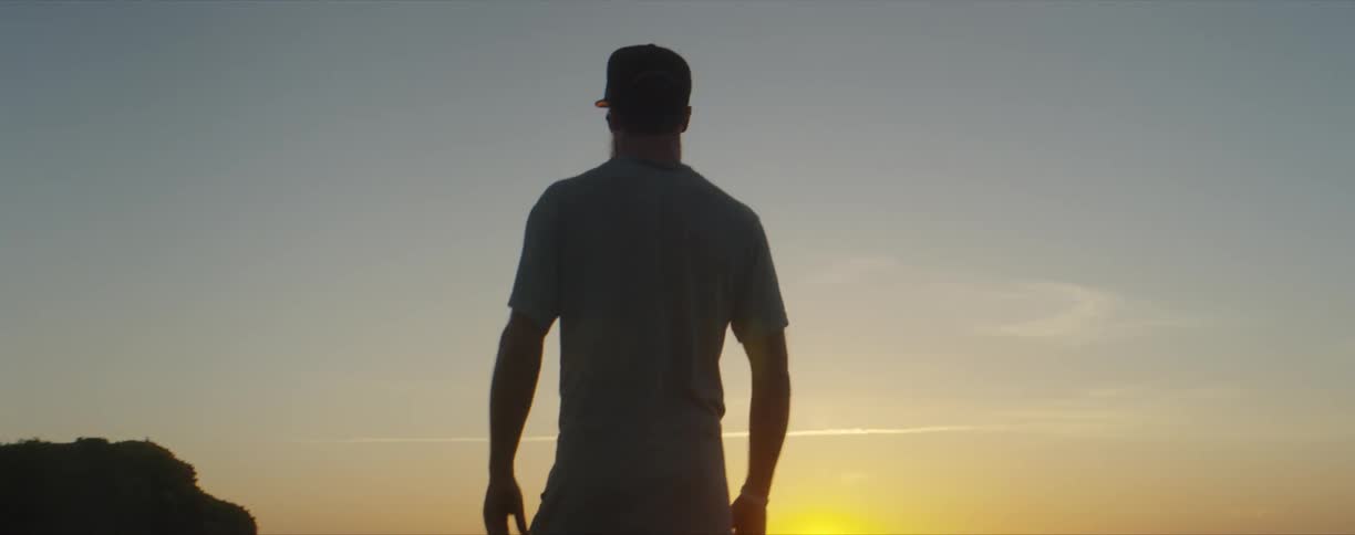 Man wearing a hat walks towards a lake as the sun sets