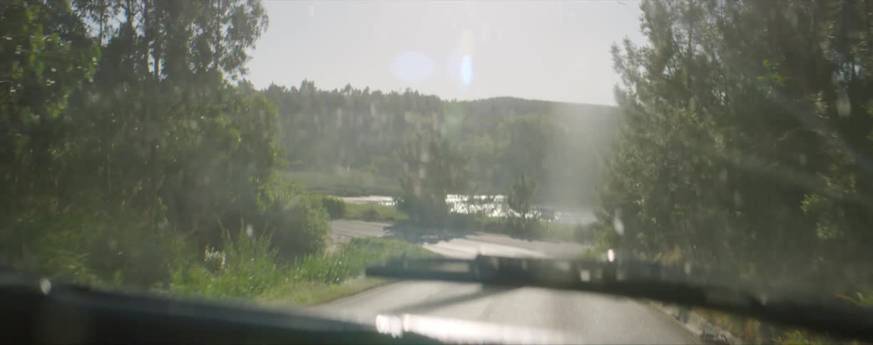 Driving down a road with trees on both sides a car lake and hill is in the distance