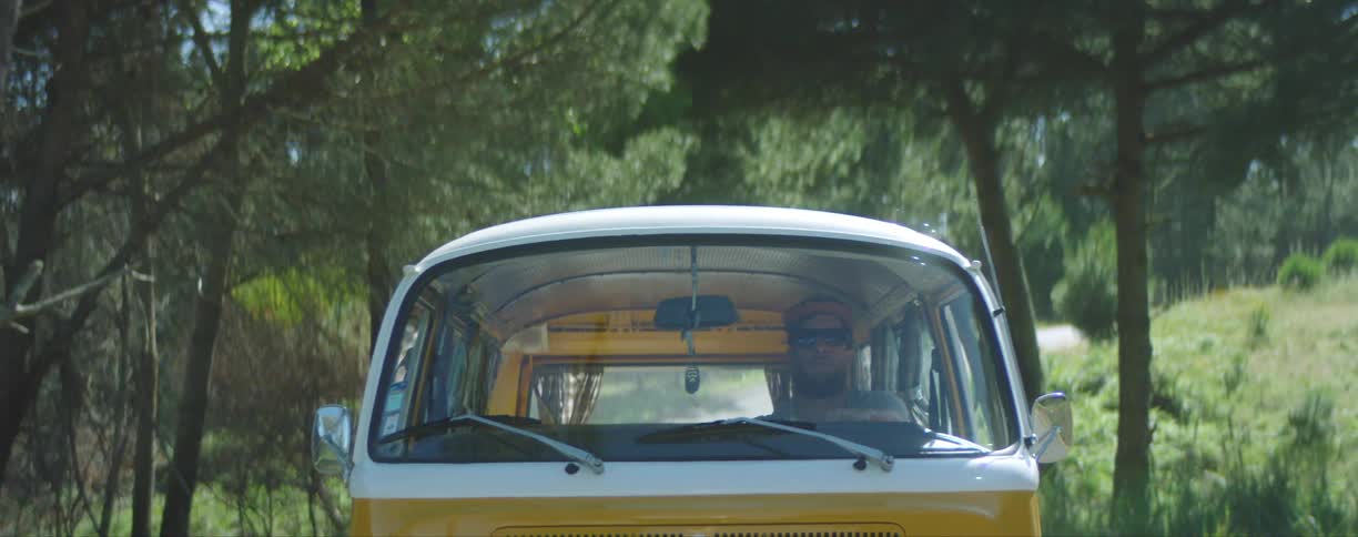 A man wearing sunglasses and a hat drives a yellow and white bus on a rural road