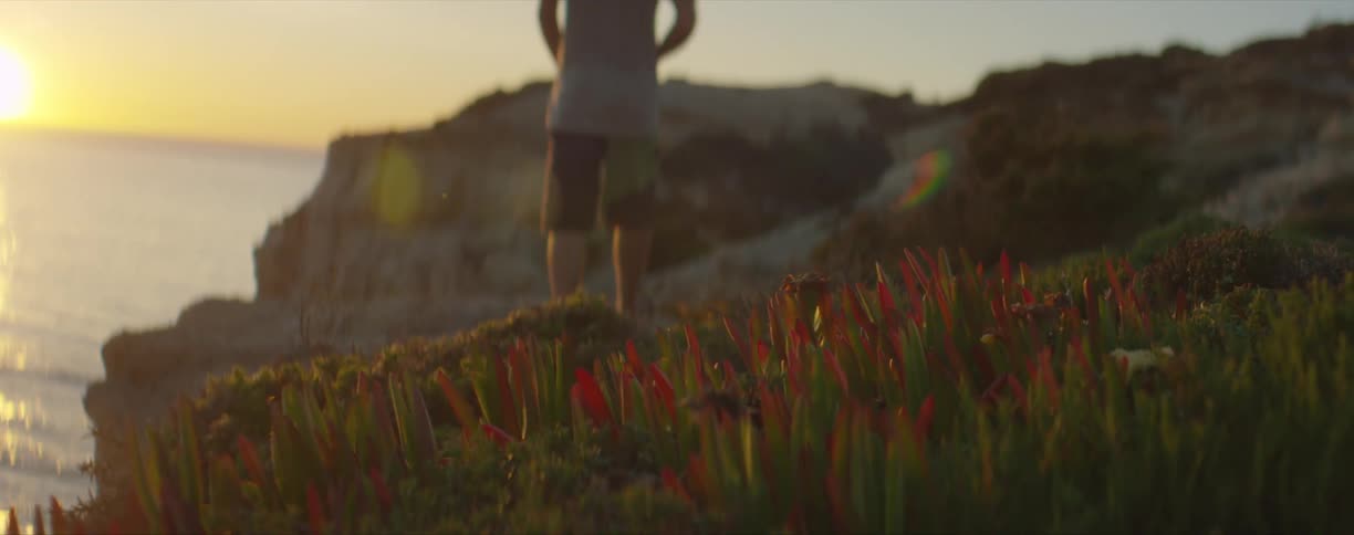 A man walks along a cliff on the shore and stops and looks at the sun setting over the water