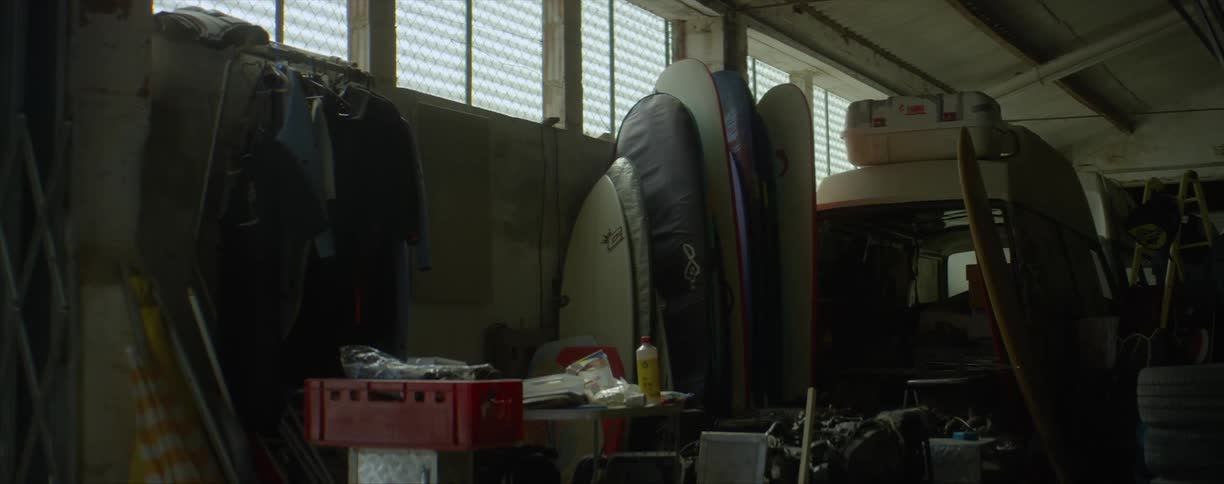 Surfboards line up against the wall of a business storage room that is filled with tools and old tires