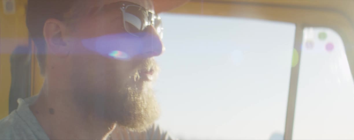 A man with a hat and sunglasses in a vehicle with the sky and clouds in the background