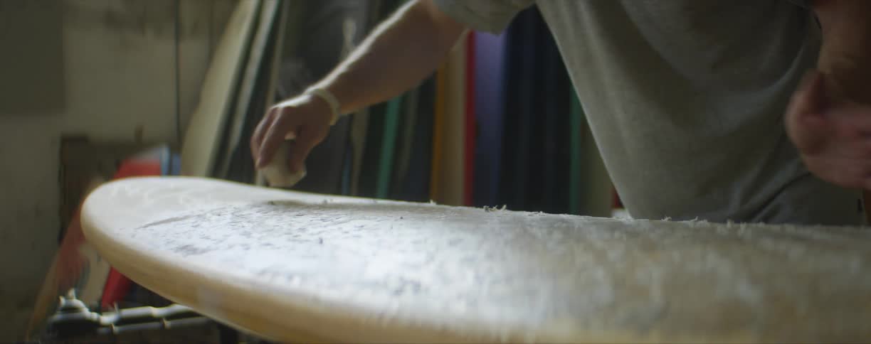 A man places wax on a surf board and wipes it with his hand while standing indoors