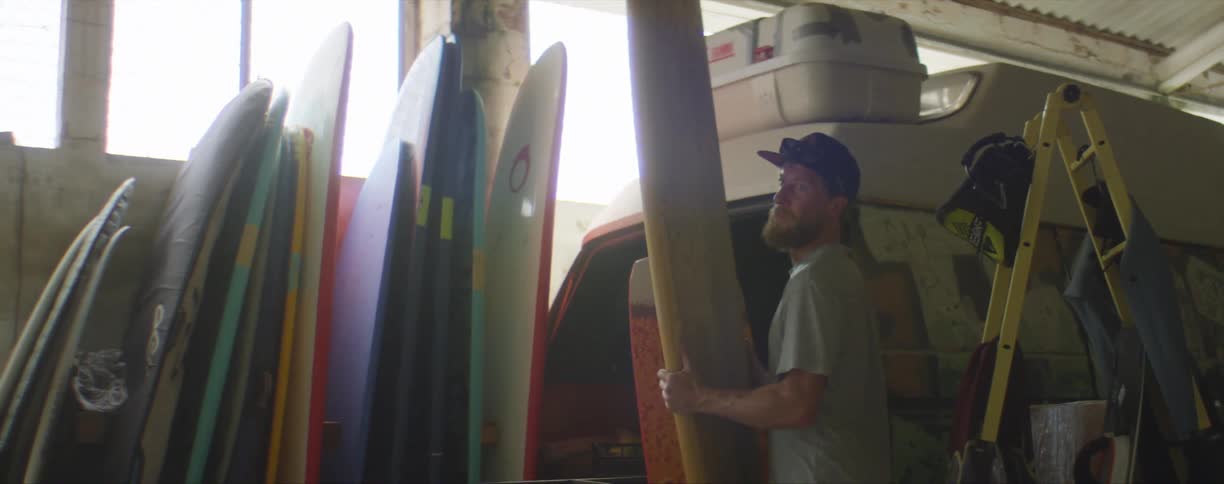 A caucasian man grabs a surfboard to put on a table with other surfboards and vans in a garage