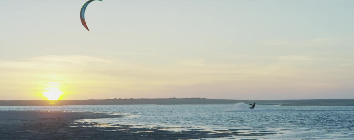 A person practicing kitesurf on the ocean next to the beach at sunset