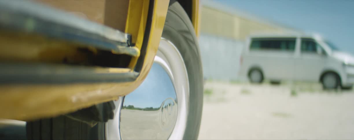 A yellow vehicle with shiny chrome rims drives out of a gravel parking lot where other vehicles and a semi sit far apart