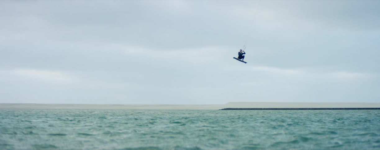 A wakeboarder is send airborne by a wave as they are pulled along the ocean on a cloudy day
