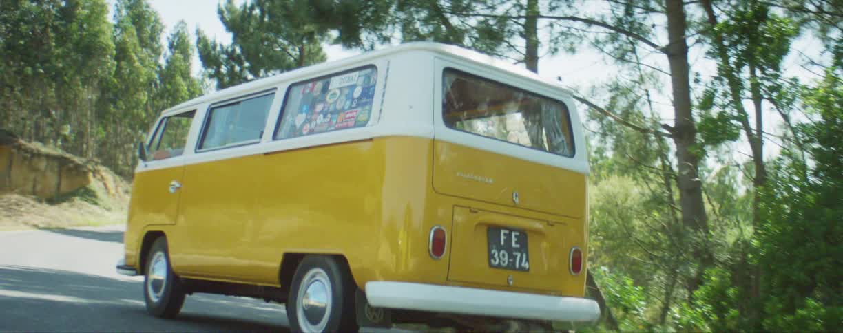 A yellow and white minibus drives on a rural road surrounded by trees
