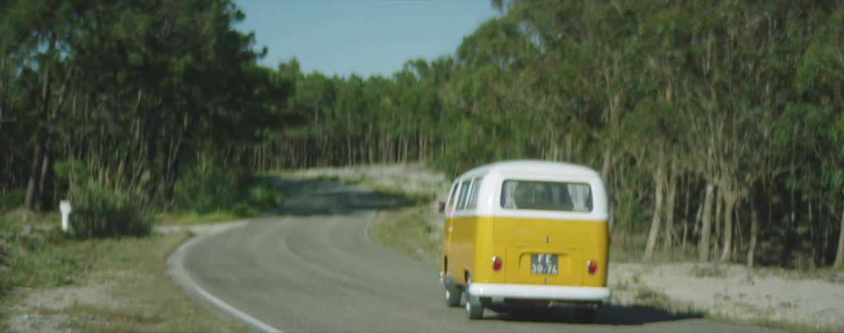 An old yellow volkswagen van driving down a winding road with trees on each side