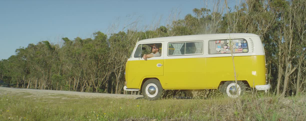 A man wearing sunglasses and a baseball hat sits in a parked yellow minibus looking out of the window