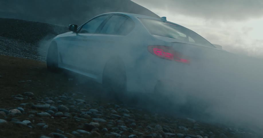 Smoke billows from a car parked in the gravel of a field in the countryside on a cloudy day