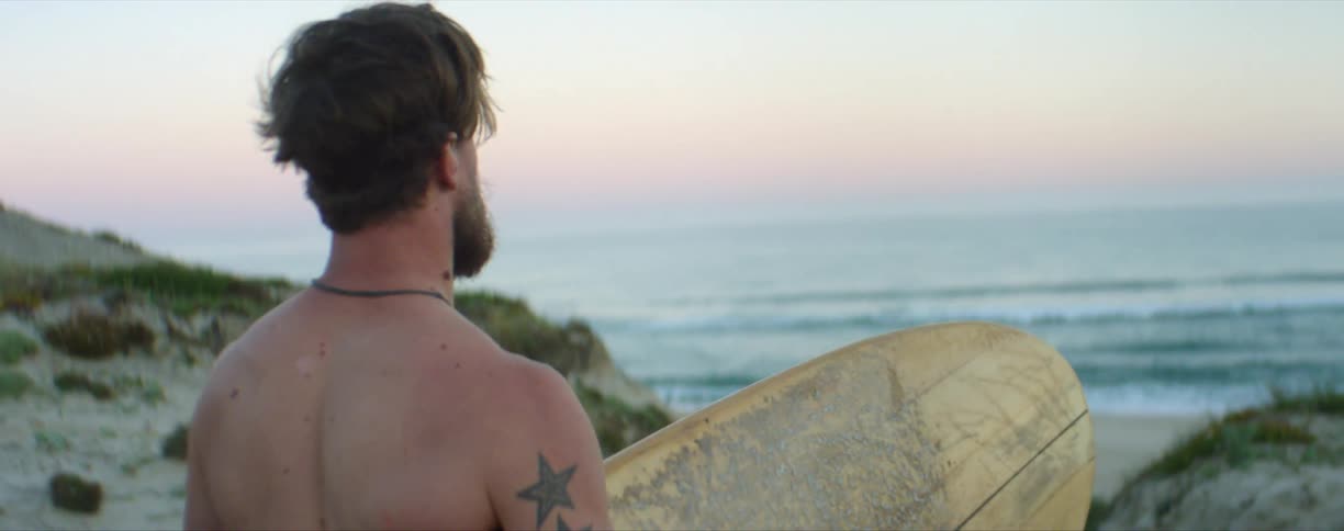 Bearded tattoed man carries a surfboard to the beach at sunset