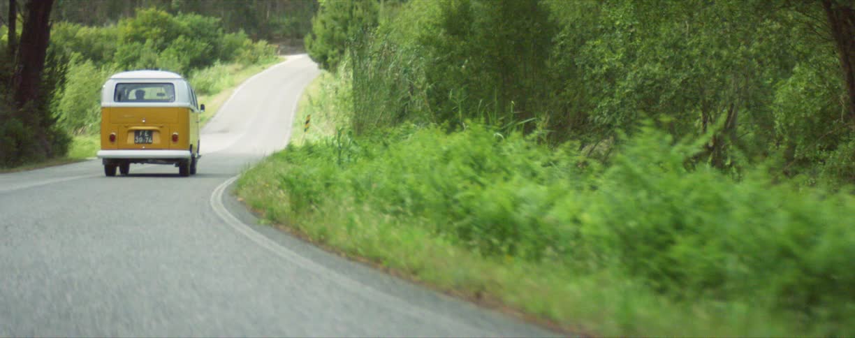A yellow and white van drives on a paved road in a rural area with trees and greeney