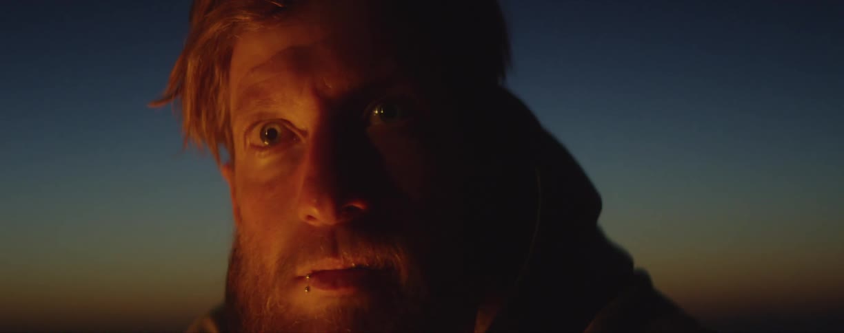 A redheaded man sits with his eyes wide open as light from a nearby fire flashes across his face