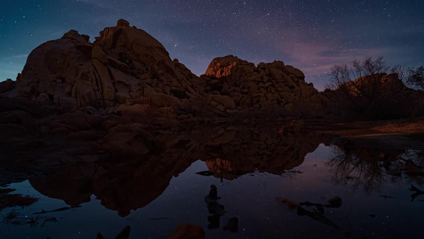 Clouds passes overhead and stars appear as night falls over rocky mountains near reflective water