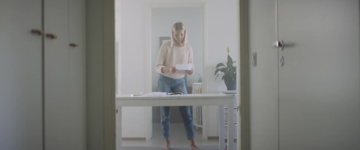 A barefoot woman organizes papers on a desk as a barefoot man carrying a baby walks behind her