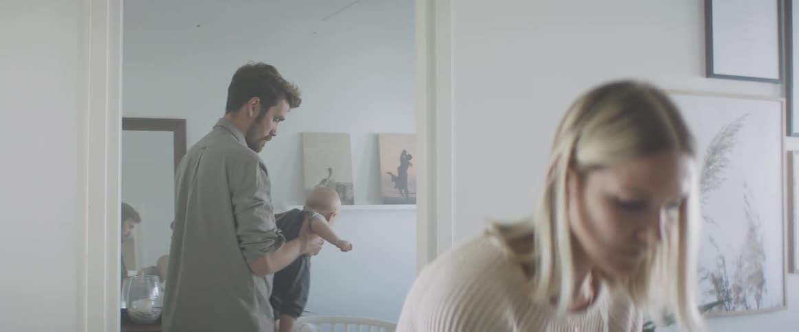 A man holding his baby lowers him to his crib and his wife is in the other room