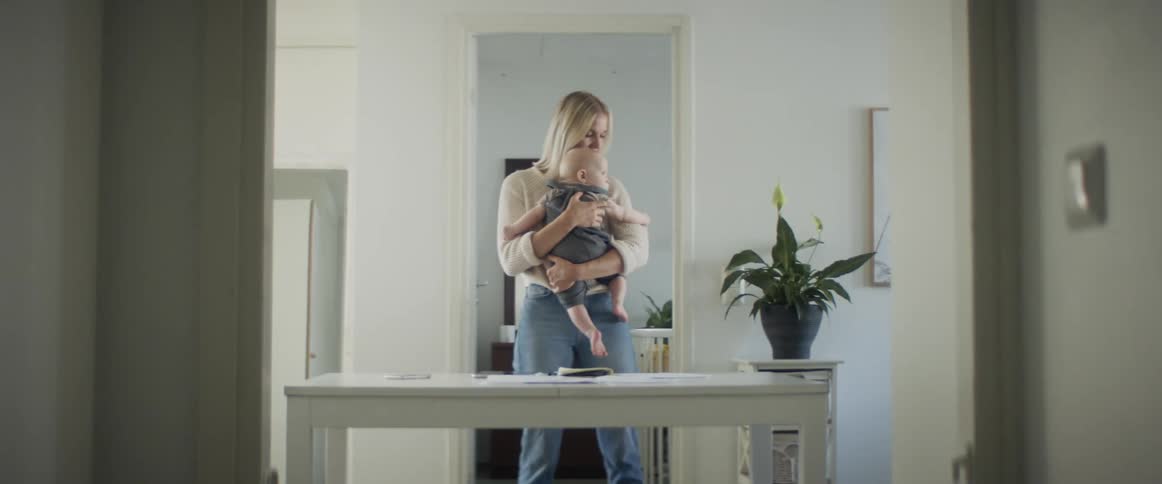 A barefoot woman stands behind a desk in a room while gently rocking the baby she carries in her arms