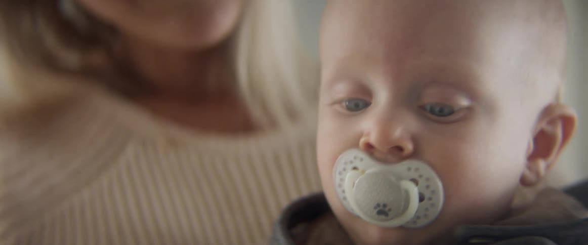 Blonde mother with a white knit sweater holds her baby son sucking a rubber nipple