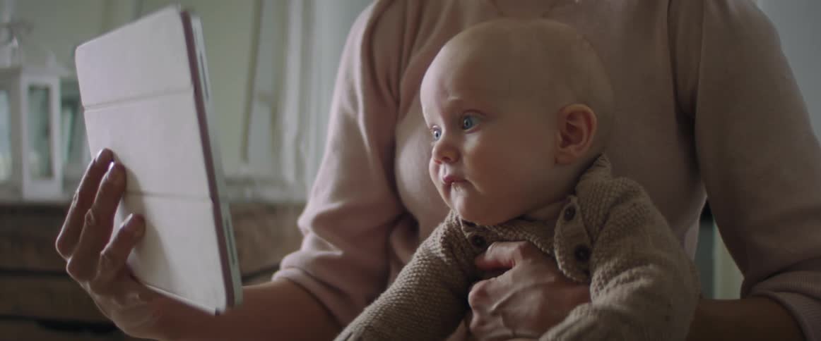 Blonde mother with a pink sweater shows a tablet to her baby son