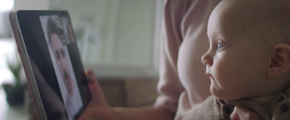 A woman holds an infant looking at a tablet of a man on a video call