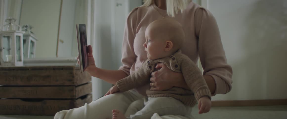 Blonde mother sitting on ground holds her baby son and shows a tablet