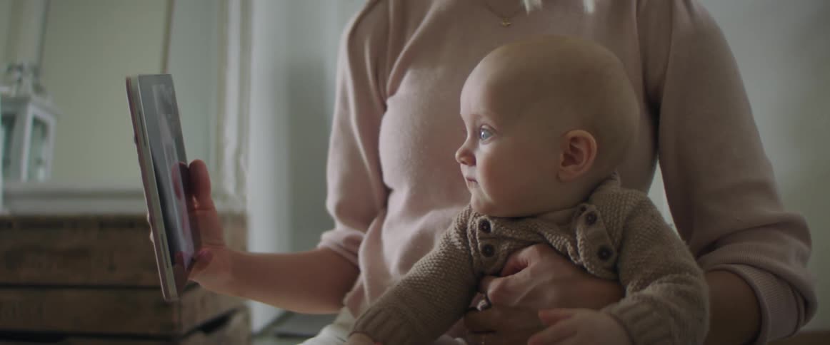 Woman sitting on the ground with a pink sweater holds her baby son and shows a tablet to the baby