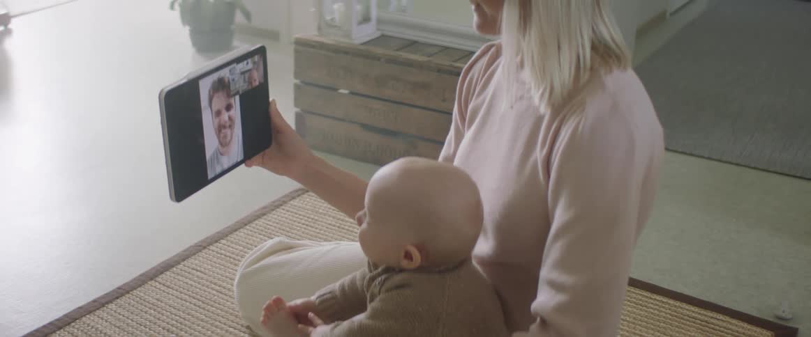 A woman sits on a rug in her house and holds her son as they video chat on a tablet with her husband
