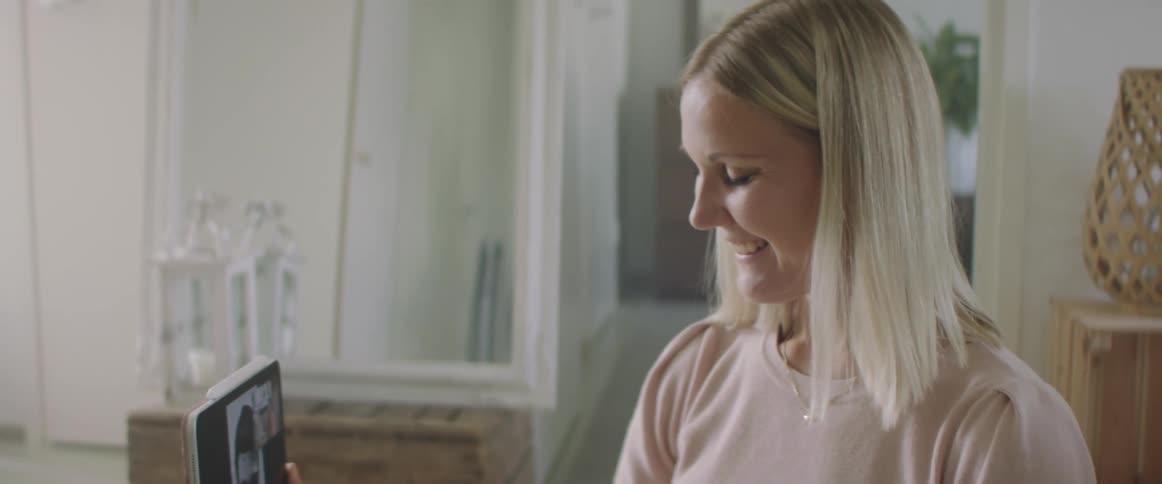 A white woman with blonde hair and a pink sweater is sitting on the floor with her male baby in her lap watching a tablet that she is holding