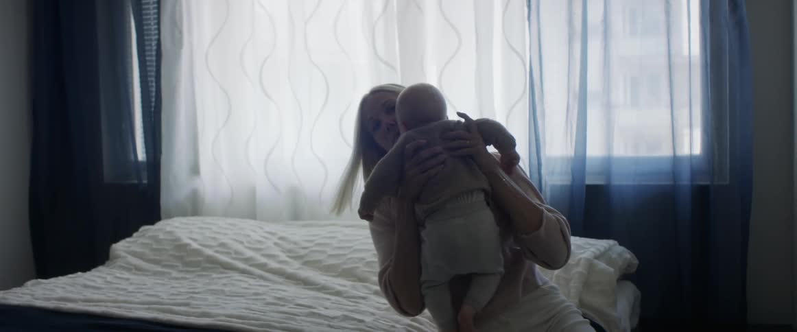 A woman picking up a baby laying on a bed in a bedroom with a window in the background