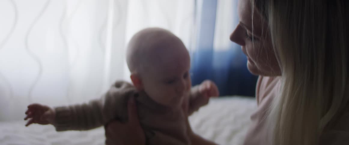 Blonde mother with a pink sweater sitting on a bed holds her baby
