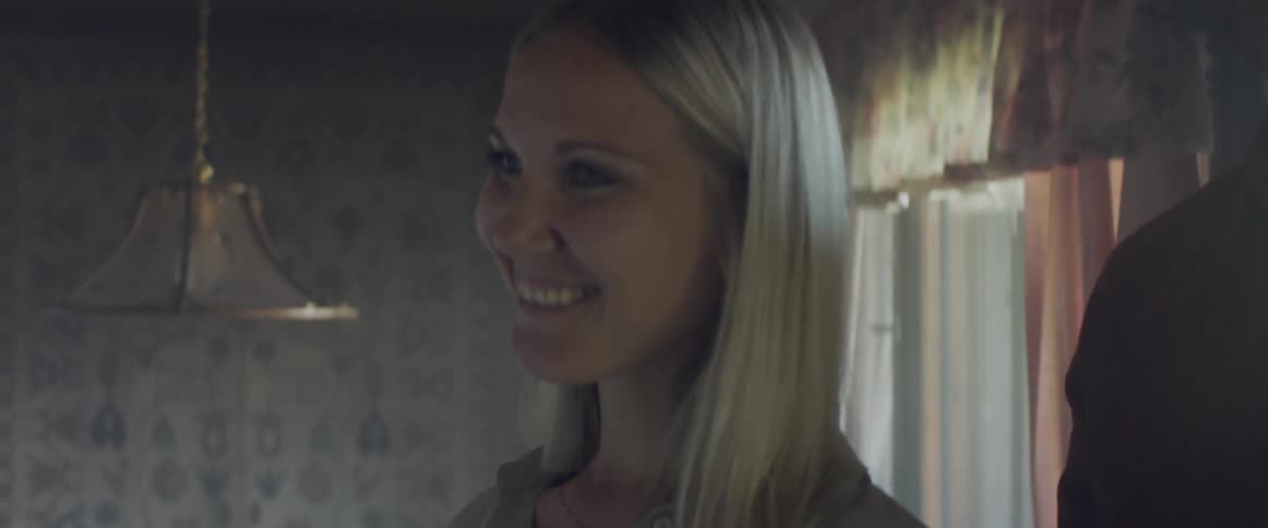 A man and woman enter a living room from the front door of a house and smile as they talk to someone outside of frame