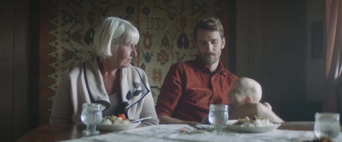 An elderly woman sitting at a table looks at her grandson who is in his dads lap and laughs