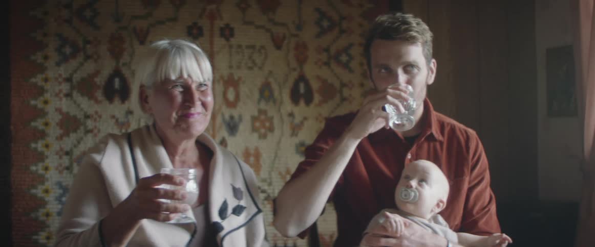 A man with a baby in his lap sits next to his mother at a table and they clink glasses together as they do a toast