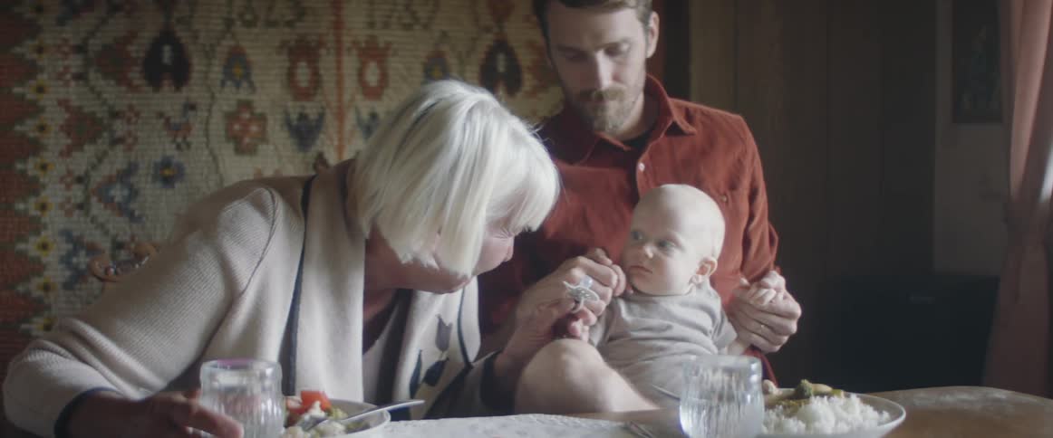 A grandmother talks to a baby on her sons knee and takes a soother out of his mouth