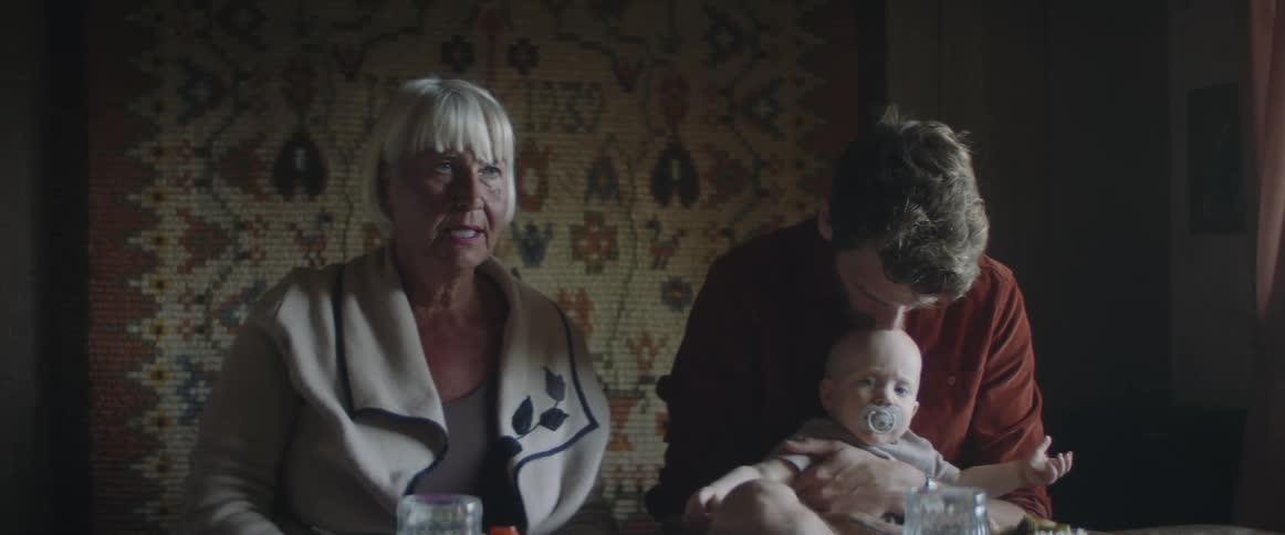 A man with a baby on his lap sits at a table next to his mother and talks while he kisses his son on the head