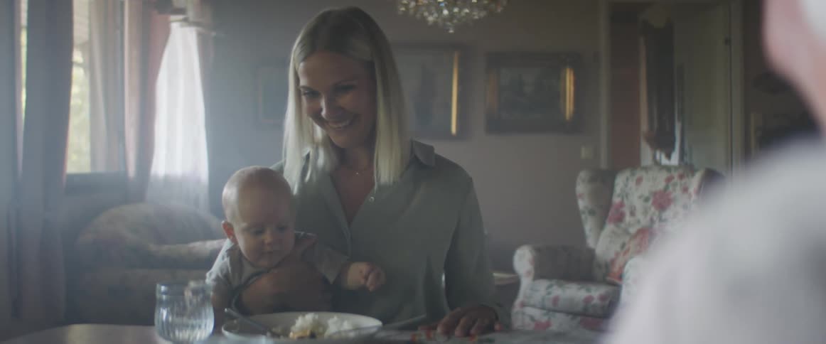 A woman sits at the table with a baby on her lap and laughs as she pulls her plate away from him