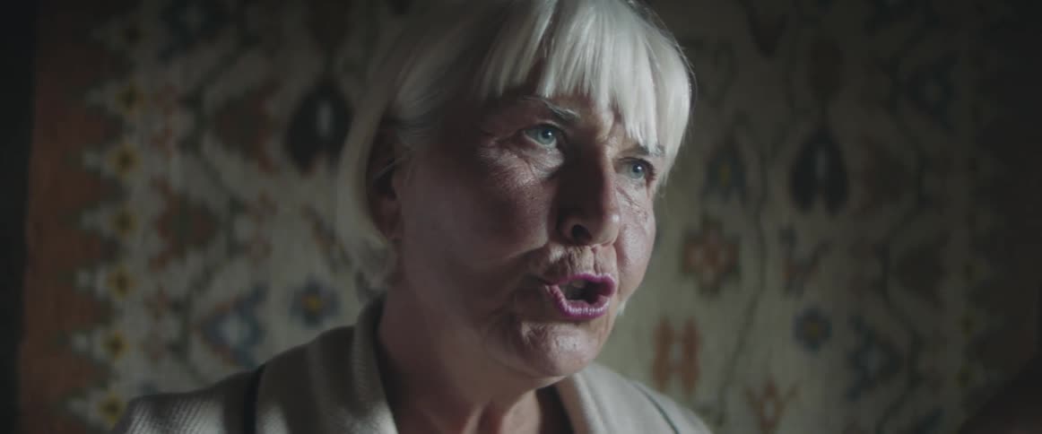 A woman with a short blonde bob haircut talks in a room with floral wallpaper