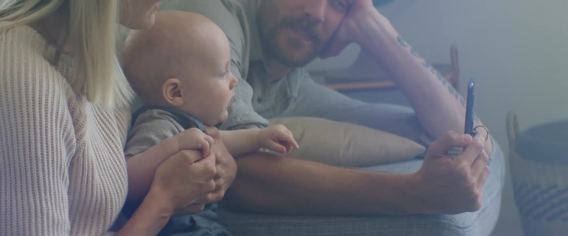 Young couple sitting in a couch at home show pictures to their baby in a phone