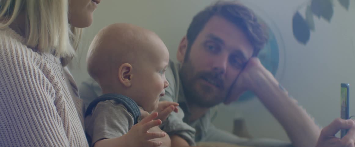 A woman holds a baby in her lap as the man sitting next to her shows the baby the screen of his smartphone