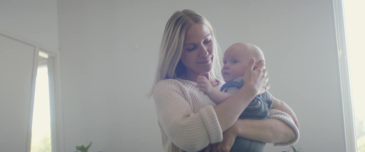 Blonde mother with a white knit sweater holds her baby and stands up in the living room