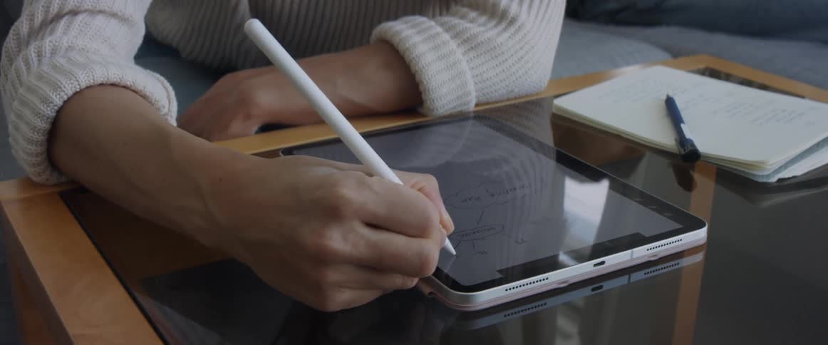 A woman sits on a couch and leans forward to draw on a tablet sitting on a coffee table