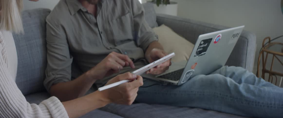 Man with a beard a shirt and jeans and blonde woman wearing a white knit shirt uses a tablet and a laptop in living room at home