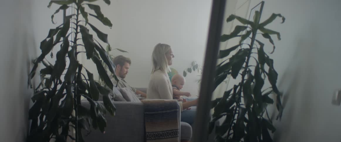 A husband and wife are shown sitting and playing with their infant son through a reflection in a full length mirror