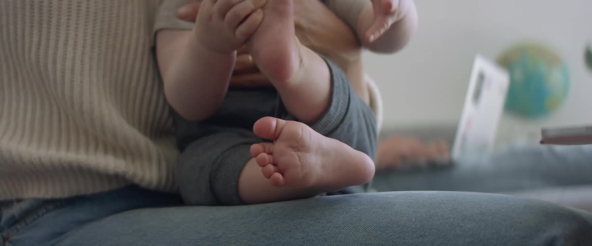 A woman sits in the living room of a house while holding a baby in her lap with one arm