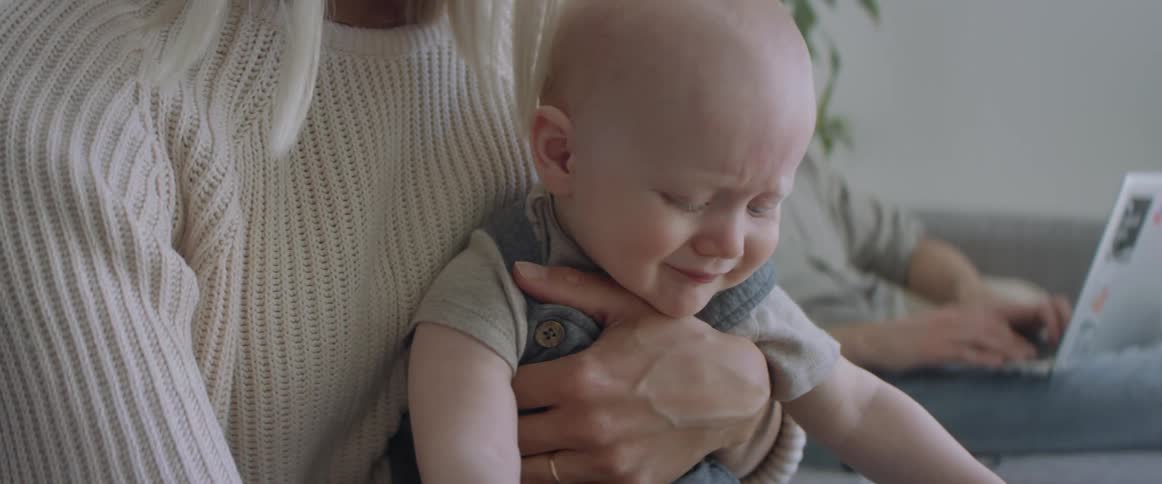 A caucasion blonde woman is holding and soothing a white infant boy in her lap while sitting on a couch that a male on a laptop is also on