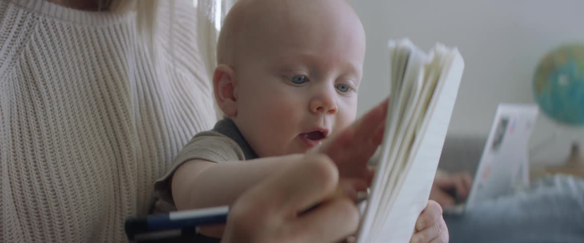 A woman tries to write in a notebook as baby being held in her arms tries to play with the paper